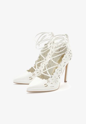 Weiße High-Heels mit spitz zulaufenden Zehen, komplizierten, ausgestanzten Blumenmustern und Schnürdetails auf einem schlanken Absatz. Weiches Ledertexture.