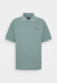 Polo vert clair à manches courtes avec une patte de boutonnage à deux boutons et un petit logo de basket noir et blanc sur la poitrine gauche.