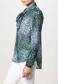 Blouse transparente avec imprimé léopard dans des nuances de vert et de bleu, dotée d'un col à nouer et de poignets froncés. Portée avec un pantalon blanc.