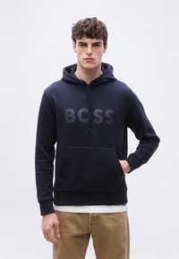 Νεαρός άνδρας που φοράει μπλε ναυτικό φούτερ με κουκούλα και μπροστινή τσέπη, με την επιγραφή "BOSS" στο στήθος, σε συνδυασμό με μπεζ παντελόνι.