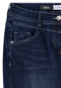 Gros plan sur un jean en denim bleu foncé avec des coutures blanches, une poche avant, des passants de ceinture, un bouton en métal et une étiquette "CECIL" sur la ceinture.