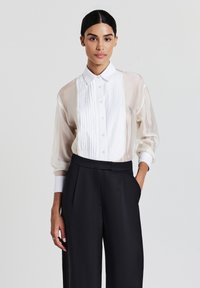 Donna con capelli scuri che indossa una camicetta bianca trasparente a maniche lunghe con parte frontale plissettata e pantaloni neri larghi a vita alta, in piedi.