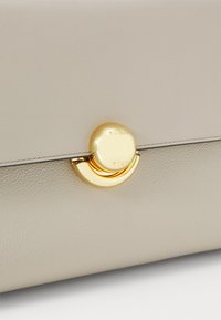 Sac en cuir beige avec surface texturée, doté d'un fermoir circulaire doré portant le nom de la marque. Design lisse avec des accents minimalistes.