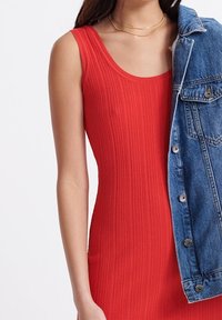 Punainen ribbikuvioinen hihaton mekko pyöreällä kaula-aukolla, yhdistettynä siniseen farkkutakkiin, jossa on hopeanväriset nappikiinnitykset ja ommeltuja yksityiskohtia.