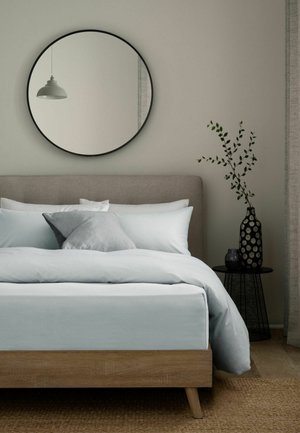 Minimalistisches Schlafzimmer mit einem Holzbett, grauen und weißen Kissen, rundem Wandspiegel, schwarzem Beistelltisch mit gemusterter Vase und grünen Zweigen.