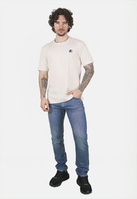 T-shirt beige a maniche corte con un piccolo logo scuro sul petto, abbinato a jeans blu e scarpe da ginnastica nere. Uomo che si presenta con sicurezza.