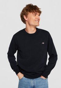Navy blue crewneck sweater made of knit fabric, featuring ribbed cuffs and hem, with a small logo on the chest. Worn with light blue jeans.