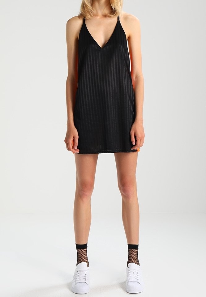 Mujer vistiendo un vestido mini negro sin mangas con finas rayas, calcetas negras de red hasta el tobillo y zapatillas blancas, de pie frente a un fondo simple.