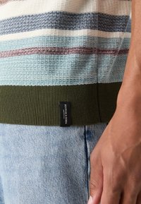 Gros plan sur l'ourlet d'un pull en maille multicolore vert à rayures avec une étiquette de la marque Scotch & Soda, porté avec un jean en denim bleu clair et une main reposant.
