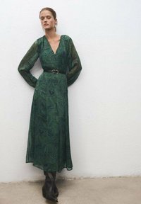 Vestido verde con estampado de cachemira, con mangas largas y transparentes, escote en V profundo, cintura ceñida con un cinturón negro, combinado con botas negras hasta la rodilla.