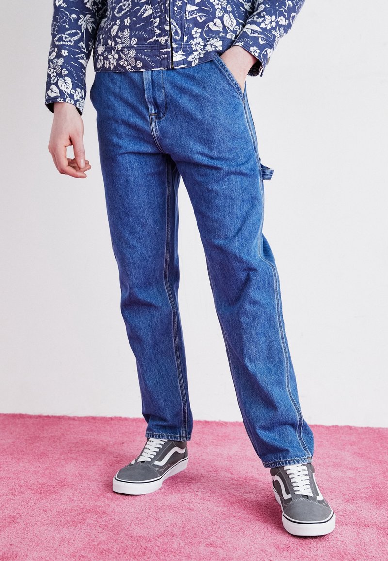 Personne portant un jean bleu, des baskets grises et blanches, et une chemise bleue à motif floral à manches longues, une main dans la poche.