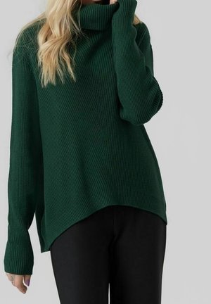 Femme portant un pull à col roulé côtelé vert foncé avec un ourlet asymétrique et des manches longues, associé à un pantalon noir.