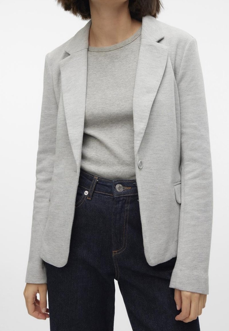 Femme portant un blazer gris clair sur un top gris assorti et un jean taille haute bleu foncé sur un fond uni.