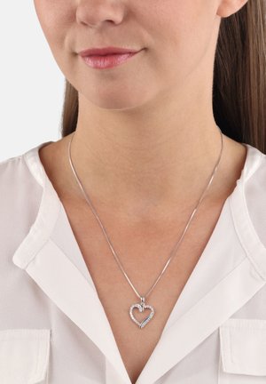 Un collier en pendentif en argent en forme de cœur avec un design entrelacé et des accents transparents, suspendu à une chaîne fine sur une blouse blanche.