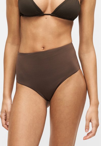 Femme portant un bas de bikini taille haute marron foncé et un haut de bikini assorti marron foncé sur un fond uni.