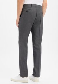 Man wearing dark gray tailored trousers and white sneakers, standing against plain white background, back view.