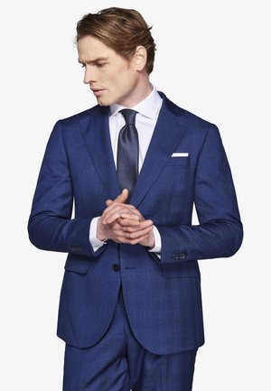 Hombre con traje azul y corbata, camisa blanca, con las manos entrelazadas, mirando hacia abajo y hacia su izquierda contra un fondo blanco liso.