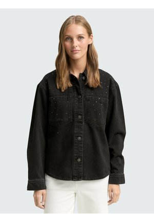 Junge Frau mit glattem schulterlangem blondem Haar trägt eine schwarze Jeansjacke mit Knöpfen, kleinen silbernen Nieten und weißen Hosen.