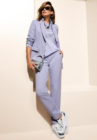 Lila blazer en bijpassende broek, gecombineerd met een licht top. Accessoires omvatten een zilveren ketting en een kleine metallic handtas. Witte sneakers.