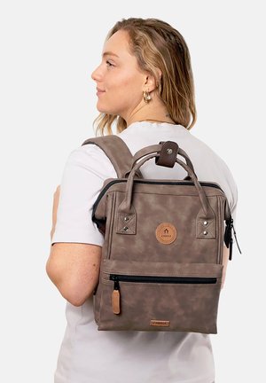 Frau mit heller Haut und blondem Haar, die ein weißes Hemd trägt und einen taupebraunen Rucksack mit schwarzem Reißverschluss und hellbraunen Lederakzenten trägt.