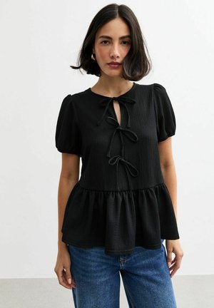 Blusa nera con texture, maniche a palloncino, apertura a occhiello e dettagli con fiocco al collo. Orlo a peplo e taglio comodo. Abbinata a jeans blu.