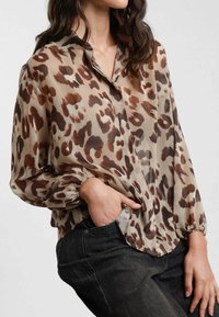 Blusa trasparente a maniche lunghe in tessuto beige con macchie di leopardato marroni, caratterizzata da un colletto e polsini arricciati. Sono indossati jeans in denim neri.