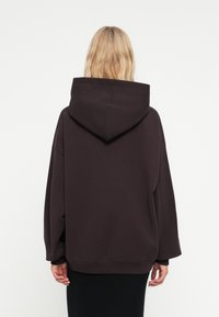 Veľká tmavohnedá mikina s veľkou kapucňou, spadnutými plecami, rebrovanými manžetami a lemom. Hladká textúra, minimalistický dizajn.