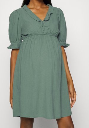 Femme en robe verte ample, longueur genou, avec des manches bouffantes et un col à volants, debout devant un fond uni.