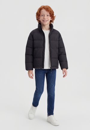 Enfant aux cheveux roux souriant, portant une doudoune noire, une chemise blanche, un jean bleu et des baskets blanches, debout devant un fond uni.