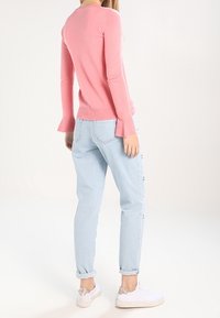 Vaaleanpunainen pitkähihainen neule flared-hihansuilla yhdistettynä vaaleansinisiin rentoihin farkkuihin ja valkoisiin tennareihin, joissa on hopeinen yksityiskohta.