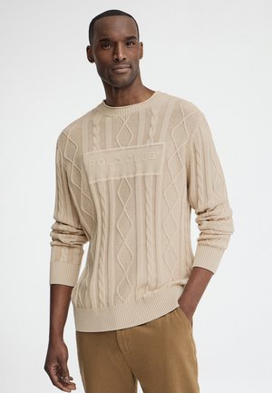 Uomo che indossa un maglione beige a trecce con il logo "Polo Club" e pantaloni color sabbia, in piedi davanti a uno sfondo chiaro uniforme.