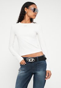 Fehér hosszú ujjú felső kis logóval, sötétkék farmerrel és nagy ezüst csattal ellátott övvel párosítva. Áttetsző napszemüveggel kiegészítve.