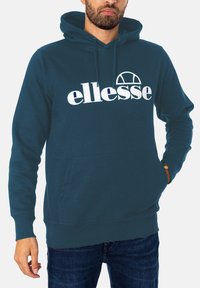Teal Kapuzenpullover mit einer Fronttasche, gerippten Bündchen und Saum sowie einem auffälligen weißen Logo, das über der Brust aufgedruckt ist. Material aus Baumwollmischung.