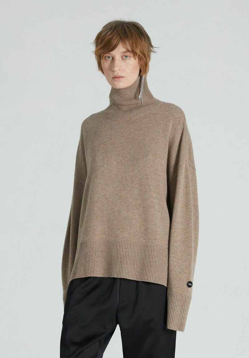 Pulover oversized bej cu guler înalt, confecționat din țesătură moale, având tiv și manșete ribbate, cu un buton de accent negru pe mânecă.