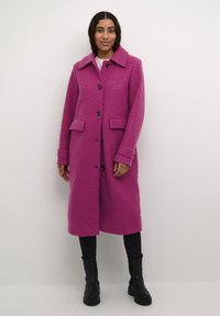 Lungo cappotto rosa imbottito con colletto, tasche frontali e chiusura a bottoni. Indossato sopra un top chiaro e abbinato a pantaloni neri e stivali.
