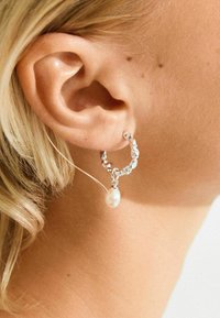 Nærbillede af en persons øre med kort blonde hår, iført en sølv hoop ørering med en hængende hvid perle.