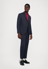 Uomo in un abito a righe navy con una polo bordeaux, calzini bianchi e mocassini neri in piedi davanti a uno sfondo bianco.