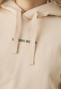 Gros plan d'une personne portant un sweat à capuche beige avec des cordons épais et un texte vert partiellement visible sur la poitrine.