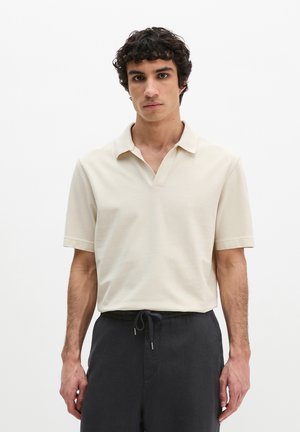 Jeune homme aux cheveux courts et bouclés portant un polo beige à manches courtes et un pantalon foncé avec cordon de serrage, debout devant un fond blanc.