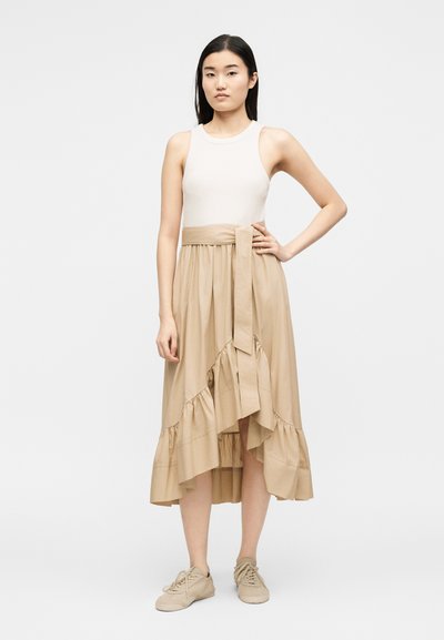 Νεαρή γυναίκα που φοράει λευκό αμάνικο τοπ, μπεζ ασύμμετρη φούστα με βολάν και δεμένη ζώνη, και μπεζ αθλητικά παπούτσια, στέκεται με το ένα χέρι στη μέση.