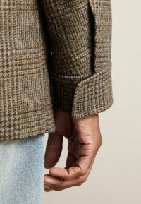 Pruunide houndstooth musteriga villane bleiser, mille mansett on tekstureeritud viimistlusega. Käsi on nähtav koos denimi detailidega, näidates vabamat istuvust ja mitteametlikku stiili.