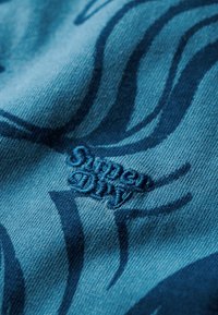 Tela azul con una textura suave que presenta patrones en espiral de color azul oscuro y un logotipo bordado de "Super Dry".