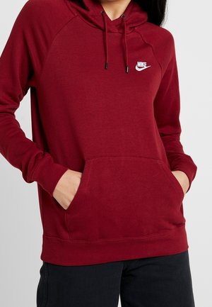 Osoba w czerwonej bluzie z kapturem Nike, z rękami w przedniej kieszeni, widoczne czarne spodnie od talii w dół.