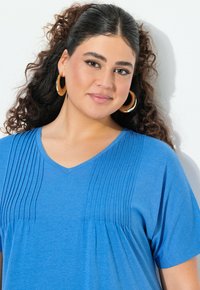 Niebieska bluzka z dekoltem w serek, z plisowanym wzorem na piersi, krótkimi rękawami i luźnym krojem. Wykonana z miękkiego, lekkiego materiału.