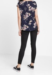 Blouse à fleurs foncées avec des manches courtes, affichant un fond bleu marine et un motif floral multicolore ; associée à un jean skinny noir et des baskets.