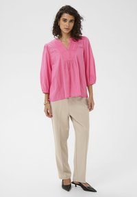 Blouse légère rose avec détails plissés, manches trois quarts, associée à un pantalon beige et des escarpins noirs à bouts pointus.