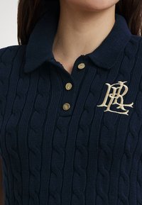 Tmavomodrá pletená polokošeľa s kablovým vzorom, s golierom, tromi zlatými gombíkmi a zlatým vyšívaným logom na ľavej strane hrude.