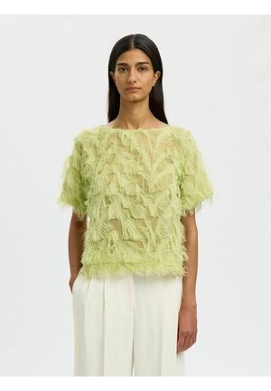 Frau trägt ein durchsichtiges, limettengrünes, strukturiertes Kurzarm-Top und hoch taillierte, lockere weiße Hosen, steht vor einem schlichten Hintergrund.