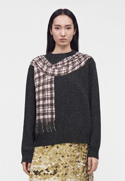 Femme aux longs cheveux foncés porte un pull gris foncé avec un motif d'écharpe à carreaux roses et noirs et une jupe en sequins dorés, face à l'avant.