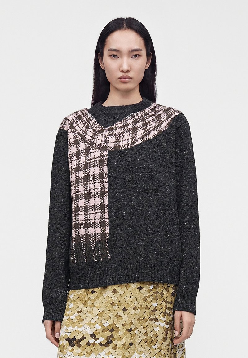 Femme aux longs cheveux foncés porte un pull gris foncé avec un motif d'écharpe à carreaux roses et noirs et une jupe en sequins dorés, face à l'avant.